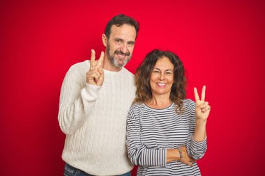 İzole kırmızı arka plan üzerinde kış kazak giyen güzel orta yaş çift zafer işareti yapıyor kameraya göz kırparak mutlu yüz ile gülümseyerek. İki numara.