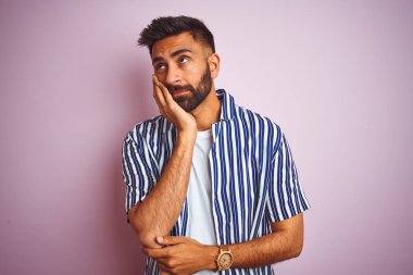 Genç yakışıklı hintli adam izole pembe arka plan üzerinde yaz çizgili gömlek giyen yorgun görünümlü ve çapraz kolları ile depresyon sorunları ile sıkılmış düşünme.