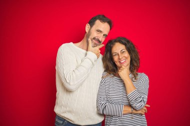 İzole kırmızı arka plan üzerinde kış kazak giyen güzel orta yaş çift çapraz kollar ve el çene kaldırdı gülümseyerek kamera emin görünümlü. Olumlu düşünme.