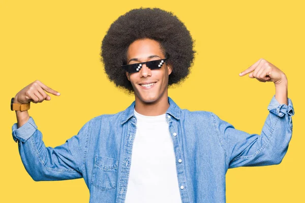 Young african american man with afro hair wearing thug life glasses looking confident with smile on face, pointing oneself with fingers proud and happy.