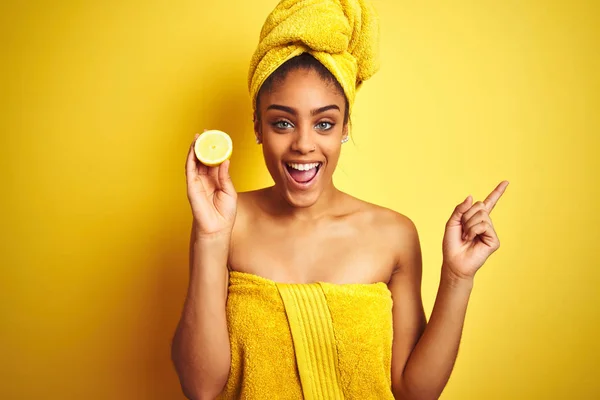 Afro woman wearing towel after shower holding slice lemon over isolated yellow background very happy pointing with hand and finger to the side