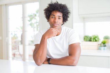 Evde oturan rahat beyaz tişört giyen genç Afro-Amerikan adam kameraya güvenerek gülümseyerek çapraz kolları ve çenesini kaldırdı. Olumlu düşünme.