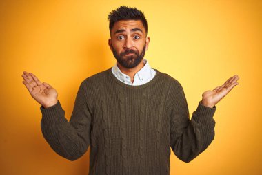 Yeşil kazak ve gömlek giyen genç hintli adam izole sarı arka plan üzerinde duran habersiz ve kolları ve elleri ile karışık ifade kaldırdı. Şüphe kavramı.