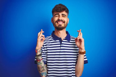 İzole mavi arka plan gesturing parmak üzerinde duran çizgili polo giyen dövme giyen genç adam umut ve gözleri kapalı gülümseyerek geçti. Şans ve batıl kavram.