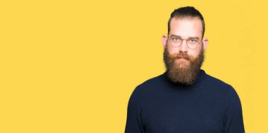 Gözlük ve balıkçı yaka kazak gevşek ciddi ifade ile yüzünde giyen genç sarışın adam. Basit ve doğal kameraya bakıyor.