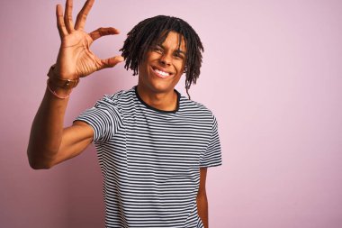 Izole pembe arka plan üzerinde duran lacivert çizgili t-shirt giyen dreadlocks giyen Afro adam gülümseyerek ve emin el parmakları görünümlü ve kamera ile küçük boyutlu işareti yapıyor gesturing. Ölçü kavramı.