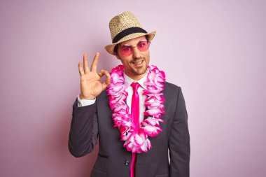 Takım elbise şapka gözlük hawaiian lei giyen genç işadamı izole pembe arka plan üzerinde parmakları ile ok işareti yapıyor, mükemmel sembolü