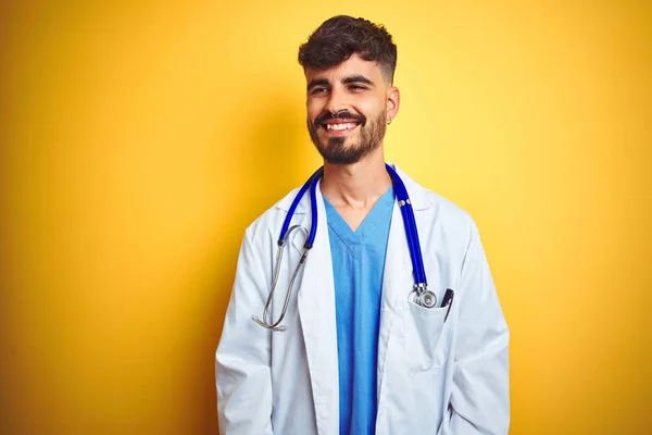 Young doctor man with tattoo wearing stethocope standing over isolated yellow background looking away to side with smile on face, natural expression. Laughing confident.