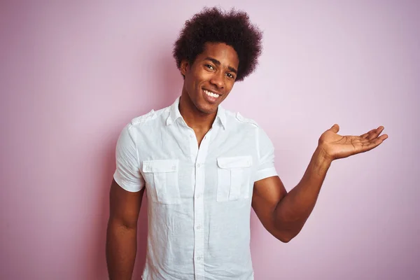 孤立したピンクの背景の上に立つ白いシャツを着たアフロヘアの若いアメリカ人男性が カメラを見て手のひらを指差して陽気な笑顔を浮かべている — ストック写真