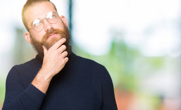 Young blond man wearing glasses and turtleneck sweater with hand on chin thinking about question, pensive expression. Smiling with thoughtful face. Doubt concept.