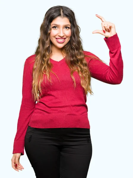 若い美しい女性の身に着けている赤いセーターの笑顔と自信を持ってカメラを見ながら指でサイズ記号をやって手でジェスチャーします 測定概念 — ストック写真