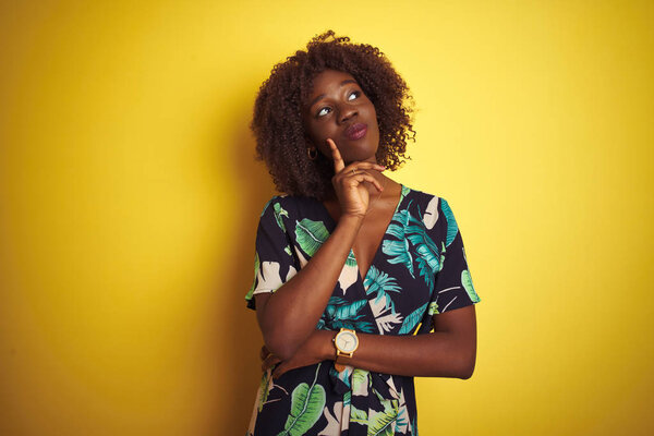 Young african afro woman wearing summer floral dress over isolated yellow background with hand on chin thinking about question, pensive expression. Smiling with thoughtful face. Doubt concept.