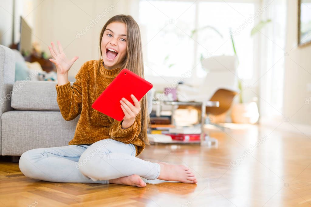 Beautiful young girl kid using digital touchpad tablet sitting on the floor very happy and excited, winner expression celebrating victory screaming with big smile and raised hands
