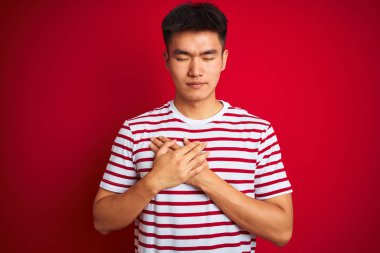 Genç asyalı Çinli adam kapalı gözleri ve yüzünde minnettar jest ile göğüs üzerinde gülümseyerek izole kırmızı arka plan üzerinde duran çizgili t-shirt giyiyor. Sağlık kavramı.