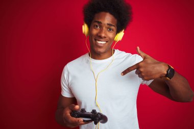 Afro oyun adam kendine parmak işaret sürpriz yüz ile izole kırmızı arka plan üzerinde joystick kulaklık kullanarak video oyunu oynuyor