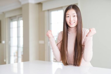 Güzel Asyalı kadın beyaz masada rahat kazak giyen sürpriz kutluyor ve kollarını kaldırdı ve açık gözleri ile başarı için şaşırttı. Kazanan kavramı.