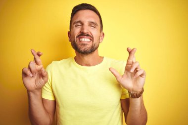 Genç yakışıklı adam sarı izole arka plan gesturing parmak üzerinde rahat sarı t-shirt giyen umut ve gözleri kapalı gülümseyerek geçti. Şans ve batıl kavram.