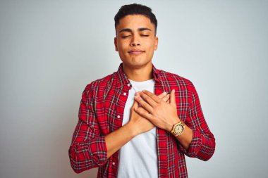 Kırmızı gömlek giyen genç brezilyalı adam kapalı gözleri ve yüzünde minnettar jest ile göğüs üzerinde gülümseyerek izole beyaz arka plan üzerinde duran. Sağlık kavramı.