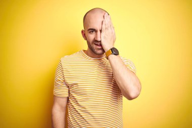Sakallı genç kel adam sarı izole arka plan üzerinde rahat çizgili t-shirt giyen el ile bir göz kapsayan, yüzünde kendine güvenen gülümseme ve sürpriz duygu.