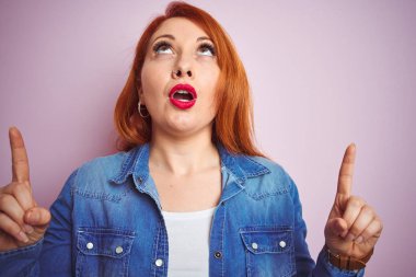 Izole pembe arka plan üzerinde duran denim gömlek giyen güzel kızıl saçlı kadın Youg şaşırmış ve baktı ve parmakları ile işaret ve kolları kaldırdı şaşırttı.