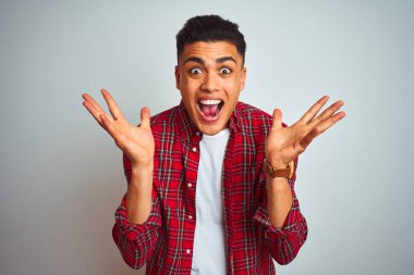 Kırmızı gömlek giyen genç brezilyalı adam izole beyaz arka plan üzerinde duran deli kutluyor ve kollarını kaldırdı ve açık gözleri heyecan çığlık ile başarı için hayran. Kazanan kavramı