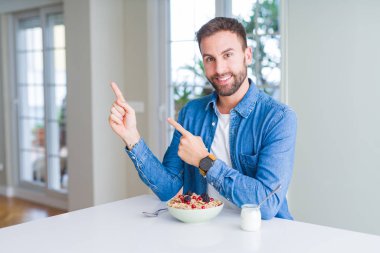 Yakışıklı adam evde kahvaltı için mısır gevreği yiyor gülümseyerek ve yan iki el ve parmakları ile işaret kamera bakıyor.