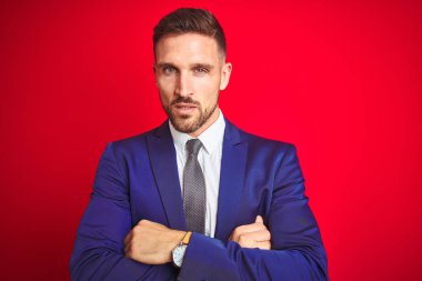 Kırmızı izole arka plan şüpheci ve sinir üzerinde genç yakışıklı iş adamı, çapraz kolları ile yüzünde onaylamayan ifade. Negatif kişi.