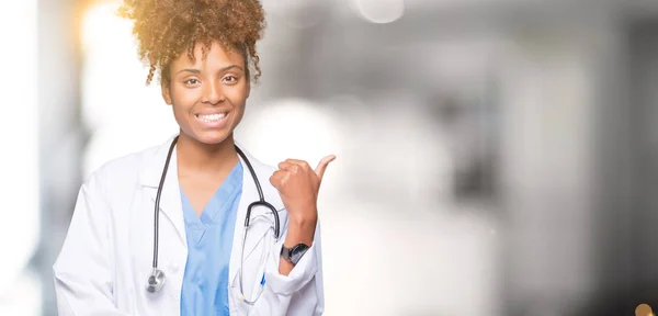 Young african american doctor woman over isolated background smiling with happy face looking and pointing to the side with thumb up.