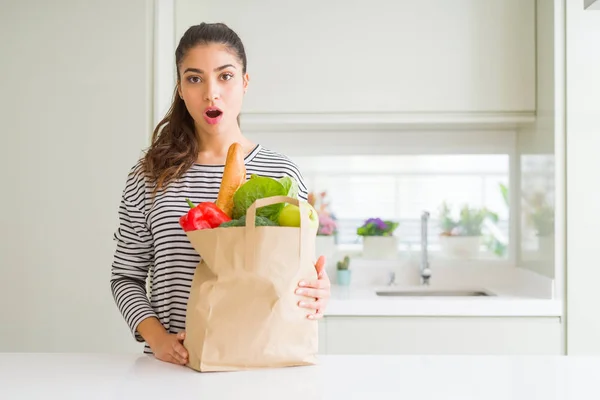 Young woman holding paper bag full of groceries scared in shock with a surprise face, afraid and excited with fear expression