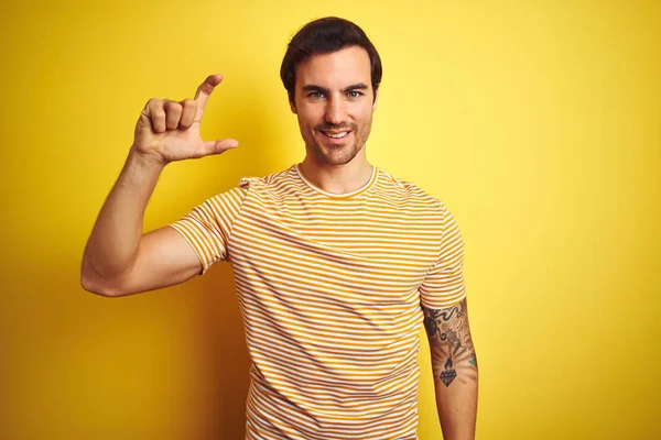年轻的英俊男子与纹身穿着条纹T恤在孤立的黄色背景微笑和自信的手势与手做小尺寸的标志用手指看和相机 测量概念 — 图库照片