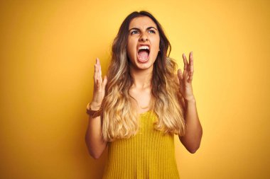 Genç güzel kadın sarı izole arka plan üzerinde t-shirt giyen deli ve deli bağırarak ve agresif ifade ve kollar kaldırdı ile bağırıyor. Hayal kırıklığı kavramı.