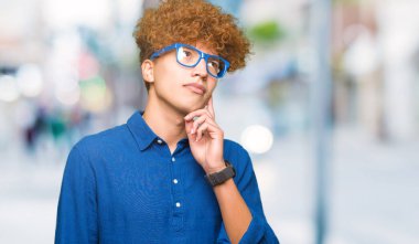 Afro saçlı genç yakışıklı adam mavi gözlük takan elleri çeneüzerinde soru, dalgın ifade. Düşünceli bir yüzle gülümsüyor. Şüphe kavramı.