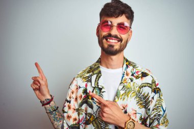 Izole beyaz arka plan üzerinde yazlık gömlek güneş gözlüğü giyen tatilde dövme ile Adam gülümseyerek ve yan iki el ve parmakları ile işaret kamera bakıyor.