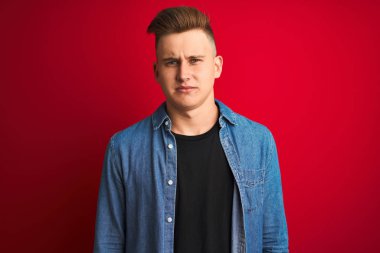Denim gömlek giyen genç yakışıklı adam izole kırmızı arka plan üzerinde ayakta depresif ve sıkıntı için endişe, kızgın ve korkmuş ağlıyor. Hüzünlü ifade.