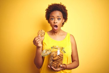 Sarı arka plan üzerinde çikolata cipsi kavanoz tutan Afrikalı Amerikalı kadın sürpriz bir yüz ile şok korkmuş, korku ifade korku ve heyecanlı