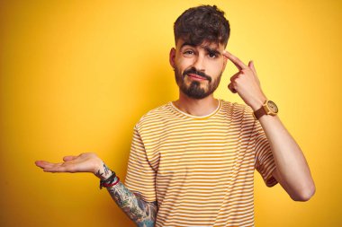 Çizgili t-shirt giyen dövme giyen genç adam izole sarı arka plan üzerinde duran karışık ve açık palmiye ile rahatsız kopya alanı gösteren ve alnına parmak işaret. Bir düşün..