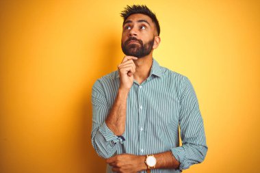 Yeşil çizgili gömlek giyen genç hintli adam, çenesi üzerinde eliyle izole sarı arka plan üzerinde duran soru, dalgın ifade düşünüyor. Düşünceli bir yüzle gülümsüyor. Şüphe kavramı.