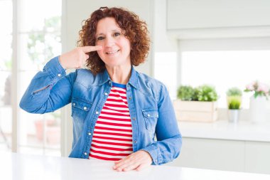 Evde kot ceket giyen kıvırcık saçlı orta yaş yaşlı kadın el parmağıyla yüz yüze ve buruna, neşeli gülümsüyor. Güzellik konsepti