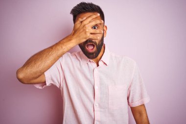 Izole pembe arka plan üzerinde duran rahat gömlek giyen genç hintli adam şok el ile yüz ve gözleri kapsayan gözetleme, utanmış ifade ile parmaklarından bakarak.