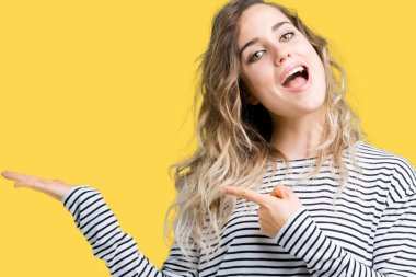 Hayret ve el ile sunulması ve parmak ile işaret ederken kameraya gülümseyen izole arka plan üzerinde çizgili süveter giyen güzel genç sarışın kadın.