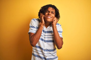 Yüksek sesle müzik sesi için rahatsız ifade ile parmakları ile kulakları kapsayan izole sarı arka plan üzerinde çizgili gömlek giyen dreadlocks ile Afro Amerikan adam. Sağır kavramı.