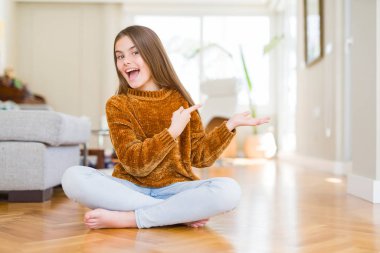 Evde yerde oturan güzel genç kız çocuk hayran ve el ile sunmak ve parmak ile işaret ederken kameraya gülümsüyor.