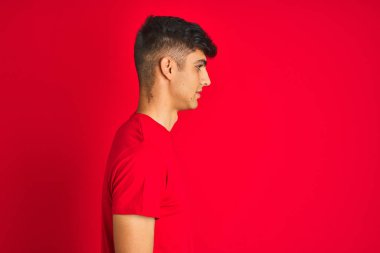 Yan bakarak izole kırmızı arka plan üzerinde duran t-shirt giyen genç hintli adam, kendine güvenen gülümseme ile doğal yüzü ile profil poz dinlenmek.