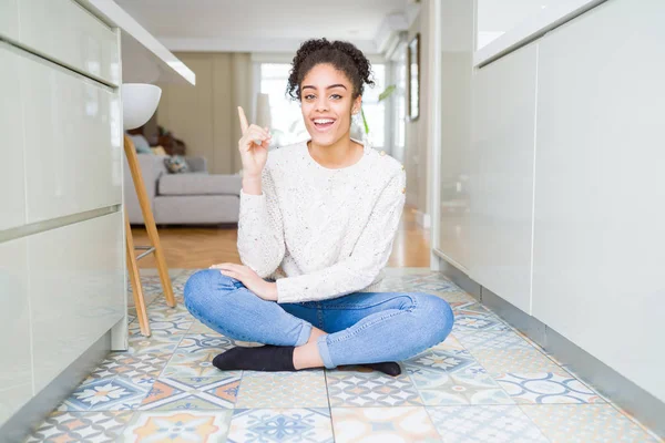 Beautiful young african american woman with afro hair sitting on the floor with a big smile on face, pointing with hand and finger to the side looking at the camera.