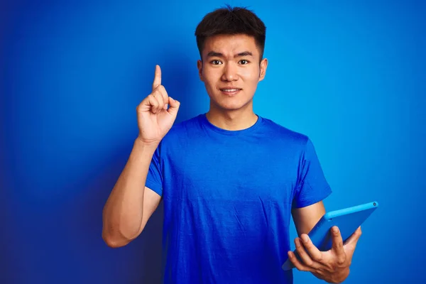 年轻的亚洲中国男子使用平板电脑站在孤立的蓝色背景惊讶与想法或问题指向手指与快乐的脸 — 图库照片