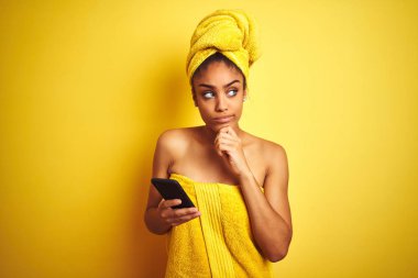 Afro kadın izole sarı arka plan üzerinde smatrphone kullanarak duş tan sonra havlu giyen ciddi yüz soru hakkında düşünme, çok karışık bir fikir