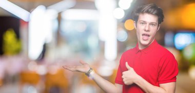 İzole arka plan gösteren palm el üzerinde kırmızı t-shirt giyen ve yukarıya, mutlu ve neşeli gülümseyen baş parmakları Tamam jest yapıyor yakışıklı delikanlı