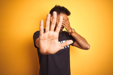 Arap Hint İspanyol koruma adam izole sarı arka plan üzerinde güvenlik üniforması giyen elleri ile gözleri kaplayan ve üzgün ve korku ifadesi ile stop jest yapıyor. Utanmış ve olumsuz kavram.