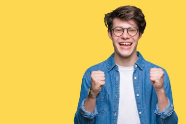 Yakışıklı delikanlı izole arka plan üzerinde kutluyor gözlük takan şaşırttı ve başarı için kaldırdı kollarını açarak hayret ve gözleri açık. Kazanan kavramı.