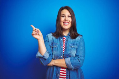 Genç güzel esmer kadın yüzünde büyük bir gülümseme ile mavi izole arka plan üzerinde rahat mavi kot ceket giyen, kameraya bakarak yan el ve parmak ile işaret.